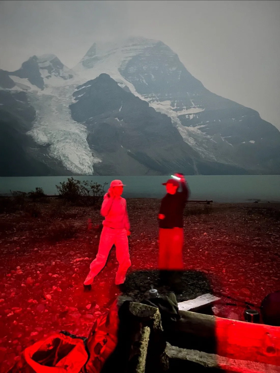 Two figures in headlamp red light standing on a stony lakeshore at night, a glaciated peak and its icefall rising grey behind the water.