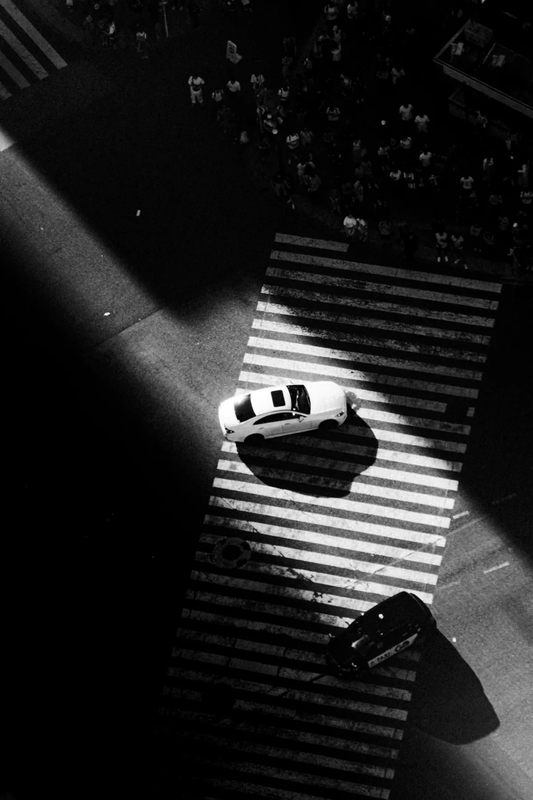 High-contrast black and white view looking straight down on a single car crossing striped pedestrian markings in raking light.