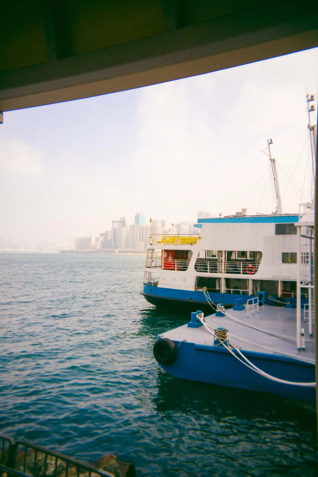 A blue and white ferry moored on calm water framed by a concrete overhang, with a hazy city skyline beyond.
