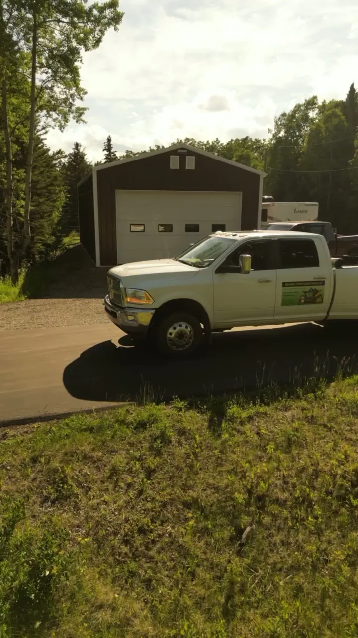 White dually pickup parked on a gravel drive in front of a dark equipment shop, summer trees and a trailer behind.