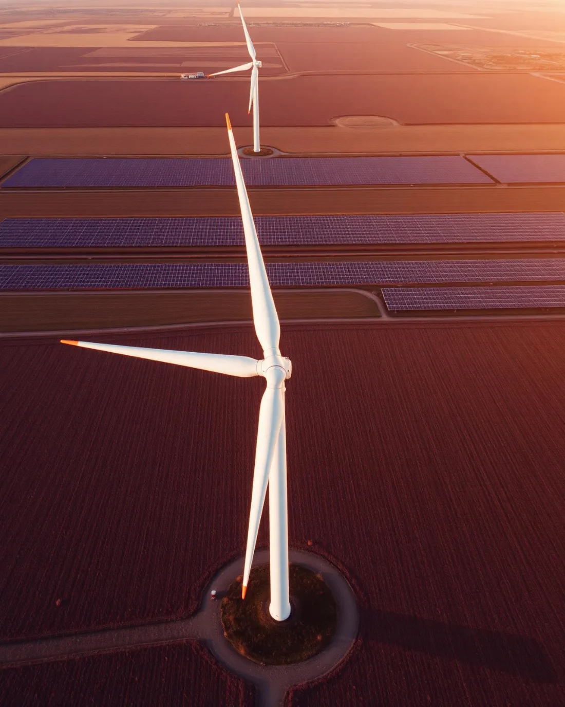 Aerial view of wind turbines standing over farmland with long rows of solar arrays catching low sunset light.