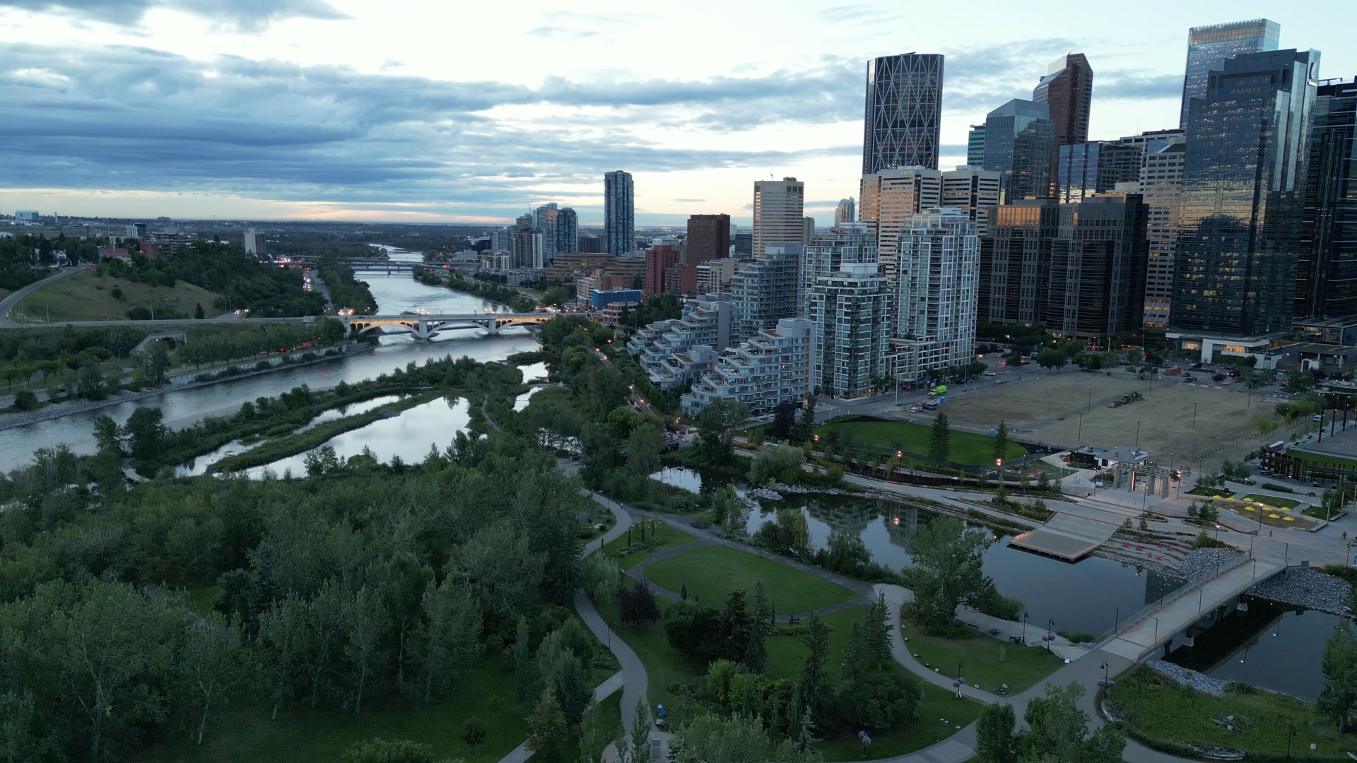 Calgary at dusk from above the Bow River: the park, the river and the downtown towers under a broken sky.